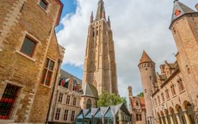 Historic courtyard in Bruges with tall Gothic church tower, brick buildings, and Gruuthuse Museum. Modern glass structure in foreground.