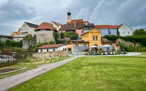 View of Schärding's historic town center with colorful baroque buildings, church tower with onion dome, and yellow Orangery building with terrace overlooking green lawn.