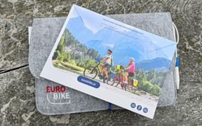 Graue Filzmappe mit Eurobike-Logo mit Reiseunterlagen und Postkarte mit Radfahrern in Berglandschaft und blauem Stift.