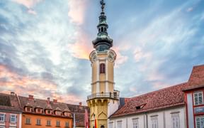 Historischer Hauptplatz in Bad Radkersburg mit barockem Glockenturm und bunten Gebäuden unter dramatischem Sonnenuntergangshimmel.