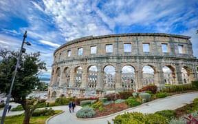Ancient Roman amphitheater in Pula, Istria with stone arches and landscaped gardens under a blue sky with white clouds.