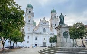White baroque St. Stephen's Cathedral in Passau with twin green copper domes and a bronze statue monument in the foreground on a square.