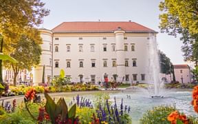 Historisches weißes Schlossgebäude in Spittal mit rotem Dach, umgeben von bunten Blumengärten und einem Brunnen im Vordergrund.