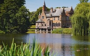 Castle Wissekerke in Flanders with red brick walls and pointed tower, surrounded by lush green trees and reflected in a tranquil pond.