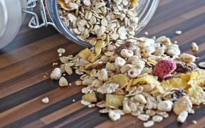 Glass jar tipped over on wooden board spilling muesli with oat flakes, dried fruits, and nuts onto striped wooden surface.