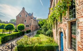 Historic brick houses with stepped gables at the Great Beguinage in Leuven. Cobblestone street with bicycles, green gardens, and ivy-covered walls.