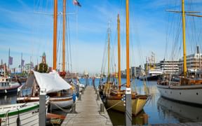 Holzsteg mit traditionellen Segelschiffen mit hohen Masten im Hafen Bremerhaven. Kirchturm und moderne Gebäude im Hintergrund unter blauem Himmel.