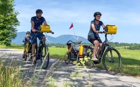 Family of three cycling on paved path with yellow panniers. Mountains and green fields visible in background under blue sky.