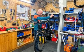 Mechanic in orange and teal jacket working on an e-bike in repair stand at Eurobike station Obertrum. Workshop has tools on wall and wheels hanging from ceiling.