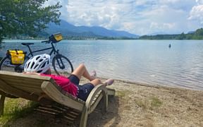 Radfahrer entspannt auf Holzliege am Ufer des Faaker Sees mit Fahrrad und gelben Taschen. Berge und blauer Himmel im Hintergrund.
