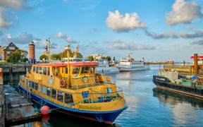 Gelb-blaues Passagierboot im alten Hafen von Cuxhaven mit rotem Leuchtturm und anderen Schiffen unter blauem Himmel mit weißen Wolken.