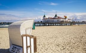 White wicker beach chair numbered 5035 on sandy beach with historic Ahlbeck pier in background. The pier features white buildings with green towers.