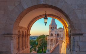 View through a stone archway at Fisherman's Bastion in Budapest showing a conical tower and city view at sunset with a hanging lantern.