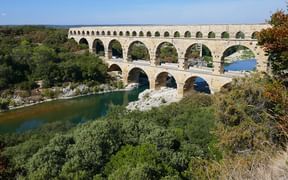 Der Pont du Gard, eine dreistöckige römische Aquäduktbrücke mit mehreren Bögen über einem Flusstal, umgeben von üppiger grüner Vegetation.