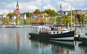Flensburg harbor with traditional fishing boat in foreground, sailboat masts, and historic town with red-brick church spire in background.