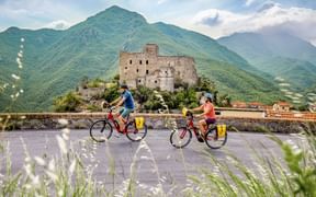 Two cyclists riding past medieval castle ruins of Castelvecchio di Rocca Barbena in Piedmont, with green mountains in the background.