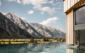 Infinity-Pool im Hotel Kristall in Leutasch mit Panoramablick auf Alpenberge, grünes Tal und modernes Holzgebäude unter blauem Himmel.