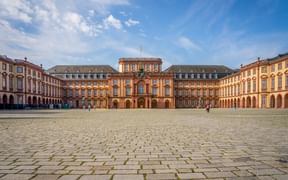 Großer Kopfsteinpflaster-Innenhof des Mannheimer Barockschlosses mit symmetrischen orange-roten Gebäuden in U-Form unter blauem Himmel.