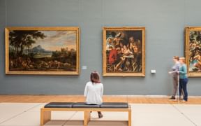Museumsgalerie mit grauen Wänden und drei großen klassischen Gemälden in Goldrahmen. Ein Besucher sitzt auf einer Bank und betrachtet die Kunst.