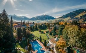 Outdoor pool at Hotel Sendlhofer's in Bad Hofgastein with garden, surrounded by trees and alpine mountains with snow-capped peaks under blue sky.