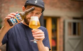 Mann hält eine belgische Bierflasche und ein Tulpenglas mit goldenem Bier vor Backsteingebäuden in Flandern.
