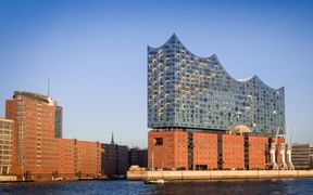 Hamburg's Elbphilharmonie with its distinctive glass wave-shaped roof on red brick base, viewed from across the Elbe River on a clear day.