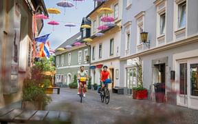 Zwei Radfahrer fahren durch eine Fußgängerzone in Villach mit bunten Schirmen, die zwischen historischen Gebäuden aufgehängt sind.