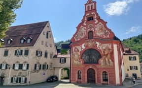 Baroque Heilig-Geist-Spitalkirche in Füssen with ornate red facade featuring religious frescoes and bell tower, surrounded by traditional buildings.