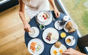 Breakfast table at Garden Hotel Reinhart with eggs, salmon, bread, yogurt, coffee and juice. Woman in white sweater arranging items.