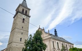Steinkirche mit hohem Glockenturm und barockem Hauptgebäude in Judenburg. Der Turm hat eine Uhr und grüne Spitze, umgeben von Bäumen.