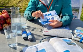 Person in türkisfarbener Vaude Radjacke an Tisch im Freien mit Reisebroschüren, Wasserkrug und Eurobike-Materialien.