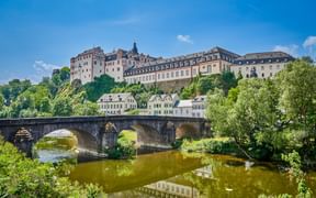 Schloss Weilburg thront auf einem Hügel über einer historischen Steinbrücke, die die Lahn überspannt, umgeben von grünen Bäumen.