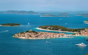 Aerial view of Primošten peninsula with terracotta roofs, church tower, and turquoise Adriatic Sea. Islands and mountains visible in background.