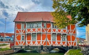 Orange half-timbered house with white timber framing and red tile roof, built over a stone bridge crossing a canal in Wismar.