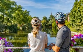 Zwei Radfahrer mit Helmen stehen an einem weißen Brückengeländer mit lila Blumen und blicken auf einen ruhigen Kanal umgeben von grünen Bäumen.