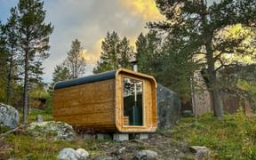 Wooden barrel sauna in Jotunheimen, Norway, surrounded by pine trees and rocks under a dramatic evening sky at Lemonsjøen Fjellstue.