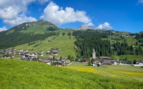 Lech am Arlberg village nestled in green Alpine valley with traditional buildings, church tower, and mountain peaks under blue sky.