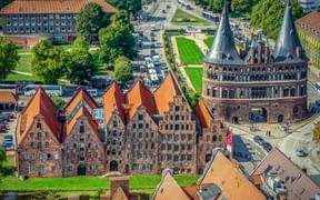 Aerial view of Lübeck's iconic Holstentor gate with twin conical towers and red brick Gothic buildings. Green lawns and streets surround the landmark.