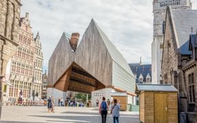 Contemporary wooden pavilion with angular roof in Ghent's historic square, surrounded by traditional Flemish buildings and a clock tower.