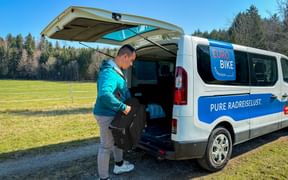 Person in türkisfarbener Jacke lädt Gepäck in weißen Eurobike-Van mit offener Hecktür. Van zeigt 'PURE RADREISLUST' Slogan in ländlicher Umgebung.