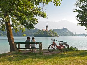 Two women taking a break from cycling on a bench on the shore of Lake Bled with a view of the island and church