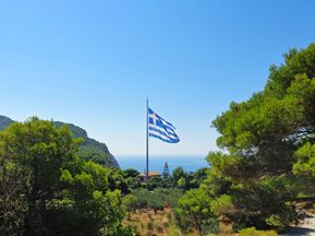 The flag of Greece, surrounded by trees and the sea in the background