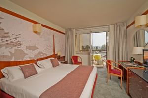 Bright hotel room with double bed, decorative wall art, balcony access, and lake view. Red and brown accents complement the neutral tones.