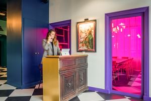 Modern hotel reception with checkered floor, wooden desk, and receptionist on phone. Purple-framed doorway reveals pink-lit room beyond.