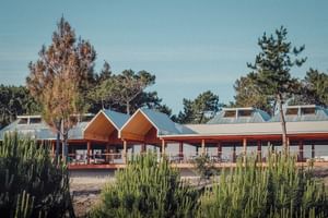 Modern hotel building with orange peaked roofs and white walls, surrounded by pine trees and green vegetation under a clear blue sky.