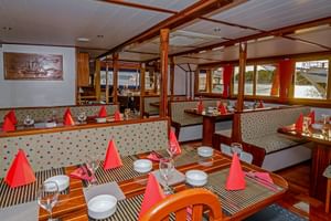 Interior of MS Macek ship restaurant with wooden tables set with red napkins, wine glasses, and striped placemats. Bench seating and windows line the walls.