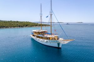 White motor sailing yacht MS Macek with two masts anchored in turquoise Adriatic Sea near green Croatian coastline under clear blue sky.