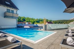 Dachterrassen-Pool im Gartenhotel Heusser Bad Dürkheim mit Sonnenliegen, weißen Handtüchern und Glasbarrieren mit Blick auf grüne Hügel.