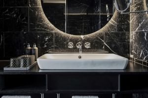 Modernes Badezimmer im The Dean München mit weißem Aufsatzwaschbecken, schwarzen Marmorwänden, beleuchtetem rundem Spiegel und Chromarmaturen.