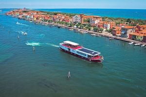 Luftaufnahme des Kreuzfahrtschiffs MS Ave Maria im blaugrünen Wasser vor Pellestrina mit bunten Ufergebäuden und hölzernen Pfählen.
